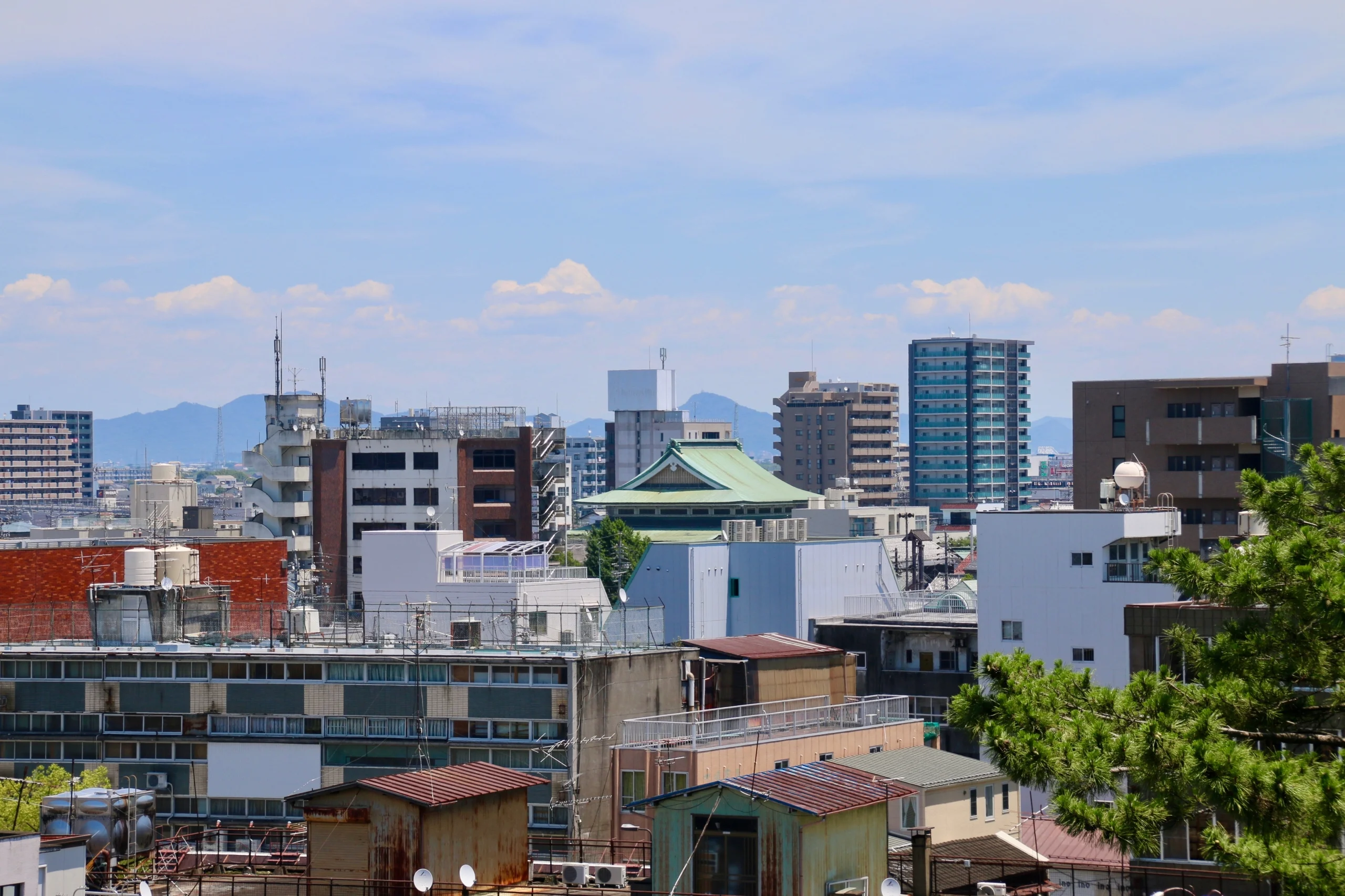 岐阜県大垣市にある大垣城から東方面を望む眺め。手前から奥にかけて、新旧さまざまなビルや住宅が密集する市街地が広がっている。街の中心には緑色の瓦屋根の建物が見え、遠景には青い山々が連なる、晴れた日の都市風景。